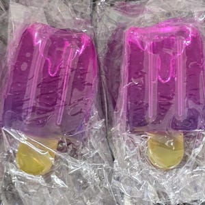 May include: Four translucent purple popsicle-shaped objects, each individually wrapped in clear plastic. The top sections feature a molded, dripping liquid design, while the bottom sections include a small, solid yellow stick handle.