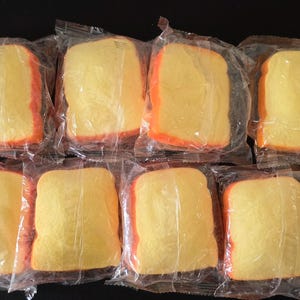 May include: Eight individual slices of yellow bread, each encased in a clear plastic wrapper. The slices have a light yellow center and a darker orange-brown crust along the edges, arranged in two rows of four against a dark background.