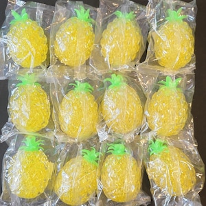 May include: A collection of twelve yellow pineapple-shaped sensory toys, each individually wrapped in a clear plastic bag. The toys feature a textured, bumpy surface and a bright green leafy top, arranged in a grid of three rows and four columns.