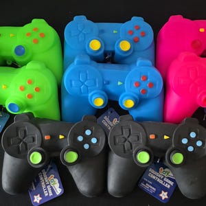 May include: A collection of seven squishy game controller toys in bright green, blue, pink, and black. Each controller features molded buttons, directional pads, and analog sticks in contrasting colors, with attached product tags labeled Toy Squishy Game Controller.