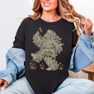 May include: A black short-sleeved t-shirt featuring a graphic print of a beige, long-haired creature resembling a Bigfoot or Yeti. The creature is depicted walking, pointing at a dragonfly, and surrounded by small, simple line-drawn flowers.