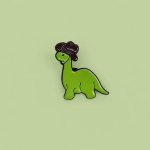 May include: A green enamel pin shaped like a long-necked dinosaur wearing a dark brown cowboy hat. The dinosaur has a smooth, glossy finish with black outlines, a small white tooth, and a rounded body profile facing toward the left side.