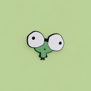 May include: A small enamel pin featuring a cartoon character with a green body and two disproportionately large white eyes with black pupils. The character has a simple, rounded shape with a small black mouth and tiny black feet at the base.