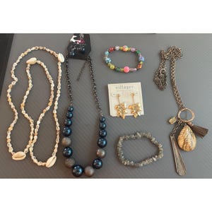 May include: A collection of jewelry including a cowrie shell necklace, a dark blue and metallic bead necklace, a multicolored bead bracelet, gold-tone leaf earrings on a Villager card, a gray stone chip bracelet, and a long chain necklace with a tassel.