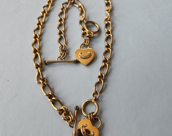 May include: Gold-toned necklace with a heart-shaped pendant featuring a smiling face design. The necklace has a toggle clasp and a chain link design. The pendant has a small diamond-like embellishment.