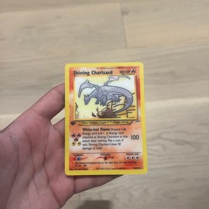Könnte beinhalten: Eine Shining Charizard Sammelkarte mit einer silberfarbenen Drachenillustration, 100 KP und der Attacke Weißglühende Flamme. Die Karte zeigt gelbe Ränder, rote und orangefarbene Hintergrundakzente sowie Text zu den Werten und Fähigkeiten des Pokémon.