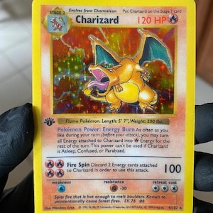 May include: A holographic Charizard trading card with a yellow border, featuring the orange dragon-like creature with blue wings. The card displays 120 HP, the Energy Burn Pokémon Power, and the Fire Spin attack, marked as a 1st Edition collectible.