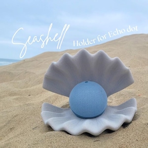 Peut inclure: Une enceinte connectée sphérique bleu clair placée à l'intérieur d'un support en forme de coquillage gris mat ouvert. Le texte "Seashell" et "Holder for Echo dot" est imprimé en blanc au-dessus de l'objet sur un fond de plage de sable.