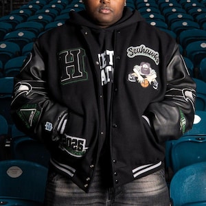 Op de afbeelding: Een zwart varsity-jack met leren mouwen, voorzien van geborduurde patches waaronder een grote letter H, het woord Seahawks, een stripfiguur met een basketbal en het jaartal 2025. Het jack heeft een drukknoopsluiting en gestreepte ribboorden.
