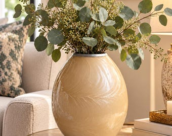 May include: A cream-coloured, oval-shaped ceramic vase with a subtle, swirling embossed feather pattern on its surface. The vase features a silver-toned rim and is filled with a large arrangement of green eucalyptus branches with small berries.