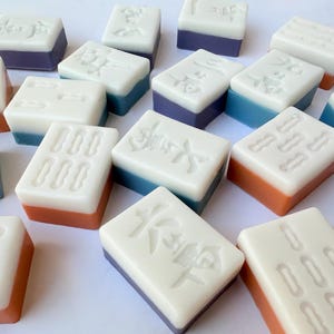 May include: A collection of rectangular mahjong tiles featuring white tops with embossed characters and symbols. The bases of the tiles are colored in various shades of orange, blue, and purple, creating a scattered arrangement on a plain surface.