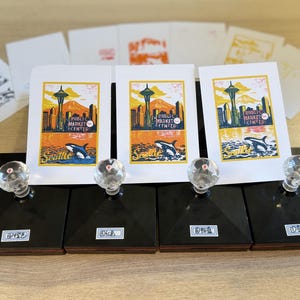 May include: Four black rectangular rubber stamps with clear crystal-like handles, each featuring a small white label with text. Behind the stamps are three identical white cards displaying a colorful Seattle print with the text Public Market Center.
