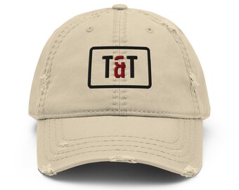 May include: A beige baseball cap with a distressed, worn-in finish. The front features a rectangular patch with a black border containing the embroidered black letters T and T with a red ampersand symbol positioned between them.