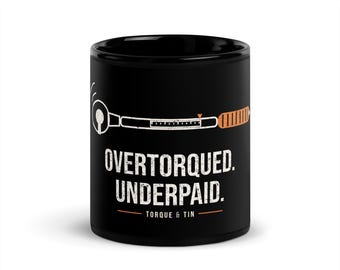 May include: A black ceramic mug featuring a white graphic of a torque wrench with an orange handle. Below the wrench, the text reads "OVERTORQUED. UNDERPAID." followed by an orange line and the text "TORQUE & TIN" in white lettering.