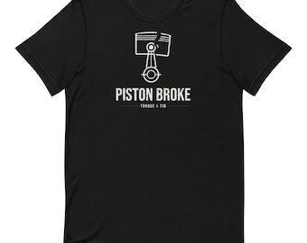 May include: A black short-sleeved t-shirt featuring a white graphic of a cracked engine piston and connecting rod. Below the graphic, the text reads PISTON BROKE in a distressed font, followed by the smaller text TORQUE & TIN.