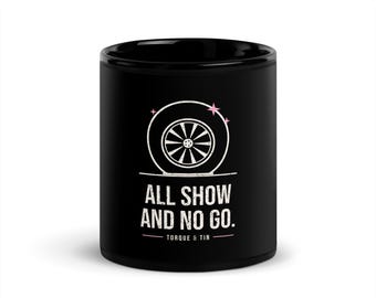 May include: A black ceramic mug featuring a white graphic of a car wheel with two small pink stars above it. Below the wheel, the text reads ALL SHOW AND NO GO. in white, followed by the text TORQUE & TIN in white with a pink horizontal line.