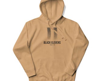 May include: A tan hooded sweatshirt featuring a graphic of two black tire tracks above the text "BLACK ELEVENS" and "TORQUE | TIN". The garment includes a front kangaroo pocket, long sleeves with ribbed cuffs, and a matching drawstring hood.