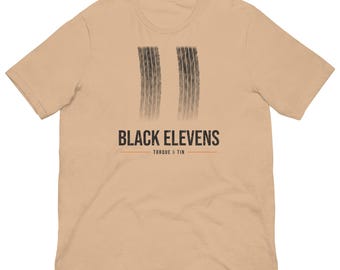 May include: A tan short-sleeve t-shirt featuring a graphic of two vertical black tyre tracks. Below the tracks, the text reads BLACK ELEVENS in a bold font, followed by a thin horizontal line and the text TORQUE & TIN in smaller letters.