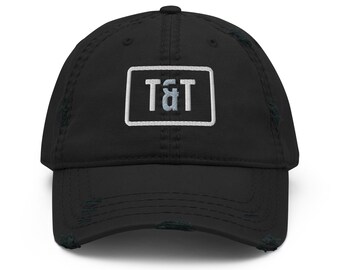 May include: A black baseball cap with a distressed, worn-in finish. The front features a rectangular white embroidered patch containing the letters T&T in a light blue and white font, centered on the crown above the curved brim.