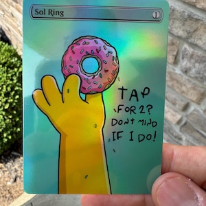 May include: A holographic trading card featuring a yellow hand holding a pink frosted donut with sprinkles. The card displays the text Sol Ring at the top and the handwritten text TAP FOR 2? DONT MIND IF I DO! on the right side against a teal background.