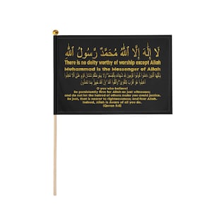 Puede incluir: Una bandera rectangular negra con texto dorado en árabe e inglés. El texto incluye la Shahada y el Corán 5:8. La bandera está unida a un mástil de madera de color claro con un pequeño remate esférico dorado en la parte superior.