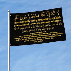 Könnte beinhalten: Eine schwarze rechteckige Flagge an einem silbernen Mast mit goldener arabischer Kalligrafie und englischem Text. Der Text enthält die Schahada und Sure 5:8: O ihr, die ihr glaubt! Seid standhaft für Allah als Zeugen in Gerechtigkeit.