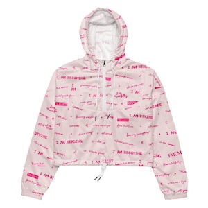 Könnte beinhalten: Ein hellrosa, bauchfreier Windbreaker mit Kapuze, halbem Reißverschluss und weißen Kordelzügen. Der Stoff ist mit einem sich wiederholenden Muster aus dunkelrosa, handschriftlichem Text bedruckt, darunter Phrasen wie I AM BECOMING und I AM DIVINE.