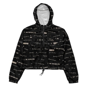 Puede incluir: Una chaqueta cortavientos negra corta con cierre de media cremallera, capucha con cordones y mangas largas. La tela presenta un patrón repetitivo de texto en blanco con frases como I AM BECOMING, I AM LIGHT y I AM DIVINE.