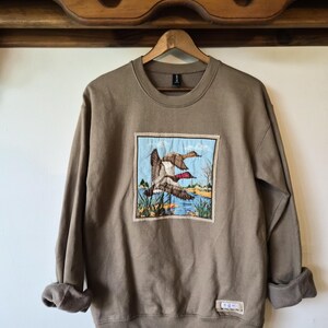 Peut inclure: Un sweat-shirt à col rond beige avec un écusson rectangulaire en tissu sur la poitrine. L'écusson présente une illustration détaillée de deux canards volant au-dessus d'un étang bleu avec des roseaux et des arbres. Une petite étiquette est cousue.