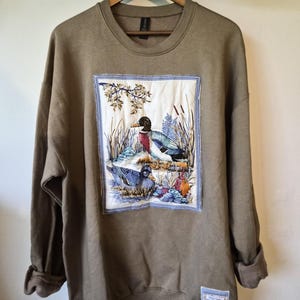 May include: A taupe crewneck sweatshirt featuring a large, rectangular, quilted fabric patch on the front. The patch displays a detailed illustration of two mallard ducks in a marshy landscape with reeds and foliage. A small rectangular label is sewn near the bottom hem.