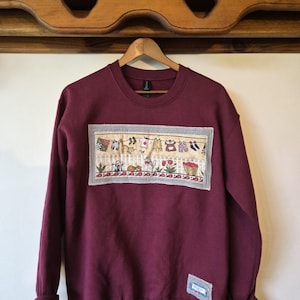 Peut inclure: Un sweat-shirt bordeaux à col rond avec une application rectangulaire en tissu sur la poitrine. L'application représente une corde à linge avec des vêtements, une clôture blanche, des chats, des fleurs et un panier d'objets rouges.