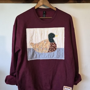 May include: A maroon long-sleeve sweatshirt featuring a large, rectangular, quilted fabric patch on the front. The patch displays a mallard duck with a dark green head, yellow beak, and a body made of floral-patterned fabric, set against a light background.