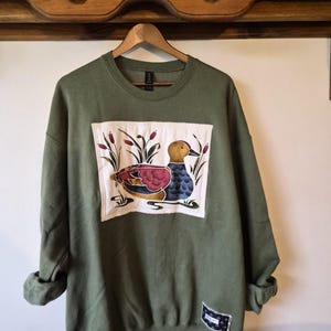 May include: An olive green crewneck sweatshirt featuring a large rectangular patch on the chest with a graphic illustration of a duck swimming among cattails. A smaller rectangular fabric label is stitched near the bottom hem.