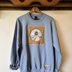 May include: A light blue crewneck sweatshirt featuring a square fabric patch on the chest. The patch displays a circular illustration of a speckled chicken against a blue sky and green field, framed by a brown dotted border with a small white tag.