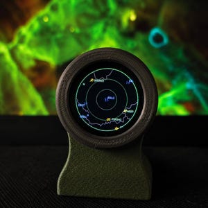 Puede incluir: Una pantalla digital circular alojada en un soporte texturizado de color verde oliva, que muestra una pantalla de radar con círculos concéntricos verdes, contornos de mapas e identificadores de vuelo como BAW665, EBLG, A82T y TOM34G.