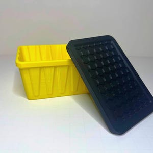 May include: A rectangular yellow storage container with vertical ribbed sides and a separate black lid featuring a raised hexagonal pattern on its surface. The container is open, and the lid is positioned at an angle next to the base.