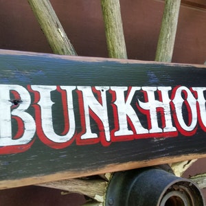 May include: A rectangular wooden sign with the word "BUNKHOUSE" in large white letters with red trim, set against a black background. The sign has a distressed, rustic appearance, mounted on a wooden wagon wheel.