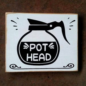 May include: White wooden sign with black text and a black and white illustration of a coffee pot. The text reads "POT HEAD".