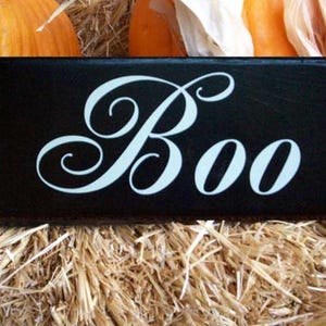 K&ouml;nnte beinhalten: Ein schwarzes Holzschild mit wei&szlig;er, geschwungener Schrift, auf dem "Boo" steht, auf einem Hintergrund aus Stroh.