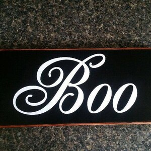 K&ouml;nnte beinhalten: Ein schwarzes Holzschild mit einer wei&szlig;en kursiv geschriebenen Aufschrift "Boo".