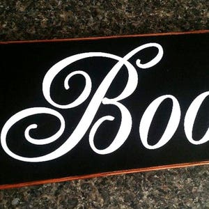 K&ouml;nnte beinhalten: Schwarzes Holzschild mit einem wei&szlig;en, kursiv geschriebenen "Boo".