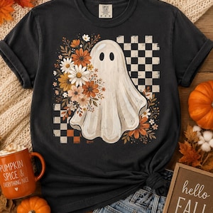 May include: A black short-sleeve t-shirt featuring a graphic print of a white ghost surrounded by orange, white, and brown flowers. The design includes a black and white checkered pattern behind the ghost. The shirt has a crew neckline and a Comfort Colors label.
