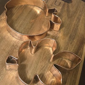 May include: Two copper-colored metal cookie cutters. One cutter is shaped like a rounded apple with a small stem at the top, and the other is shaped like a multi-lobed leaf with a pointed stem. Both items feature a metallic finish.