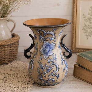 May include: A ceramic vase with a tan base color featuring a blue floral pattern and scrolling vine designs. The vase has a wide, flared rim and two dark, curved handles on opposite sides. The surface is glossy with a smooth, rounded body.