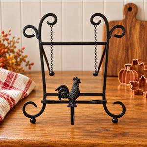 May include: A black metal display stand featuring a decorative rooster figure at the center, scrollwork feet, and two hanging chains with ball ends. The frame has a rectangular support structure with curved top accents and a sturdy base.