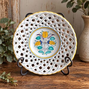 May include: A round ceramic plate featuring a white center with a hand-painted floral motif in yellow, teal, and blue. The plate has a wide, lattice-style openwork border with a scalloped yellow rim, supported by a black metal display stand.