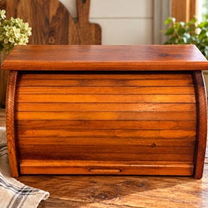 May include: A rectangular wooden bread box with a warm brown finish and a curved roll-top door. The front features horizontal slats that form the sliding mechanism, while the top is a flat, solid surface. It has a simple, functional design.