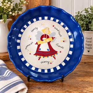 May include: A round ceramic pie dish with a deep blue scalloped rim and a white and blue checkered border. The center features a cartoon angel holding a yellow rolling pin, surrounded by the words family, traditions, friends, and sharing.