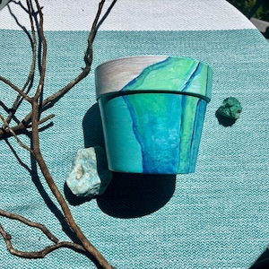 Op de afbeelding: Een taps toelopende cilindrische plantenpot met een handgeschilderd abstract ontwerp in tinten turkoois, teal en heldergroen. De bovenrand is geschilderd in een neutraal lichtgrijs, terwijl het lichaam vloeiende, gelaagde blauwe penseelstreken toont.