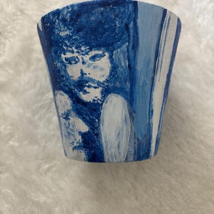 May include: A tapered cylindrical planter featuring a hand-painted design in shades of blue and white. The artwork depicts a stylized portrait of a person with textured hair, rendered in deep blue brushstrokes against a lighter blue and white background.